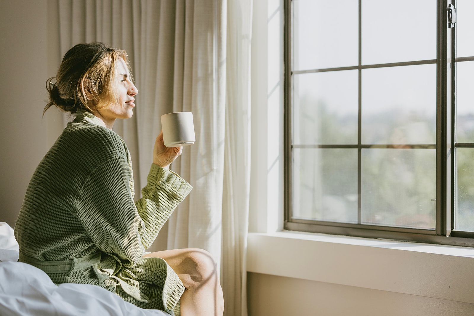 A woman in a cozy green robe enjoys a cup of coffee while gazing out of a sunlit window at Lyle DC hotel.