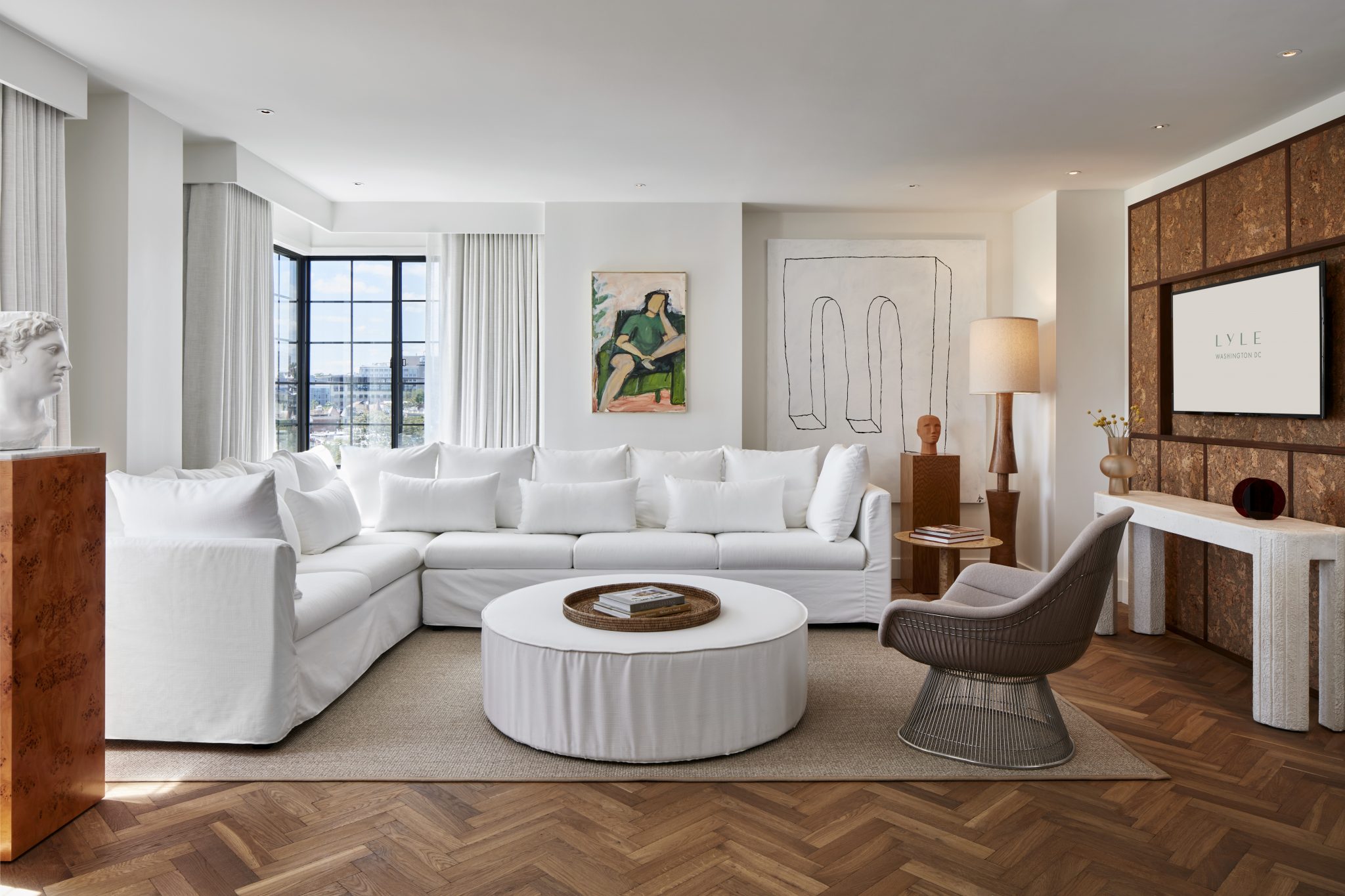 A bright and modern hotel suite at Lyle Washington DC, featuring a white sectional sofa, wood accents, and large windows.
