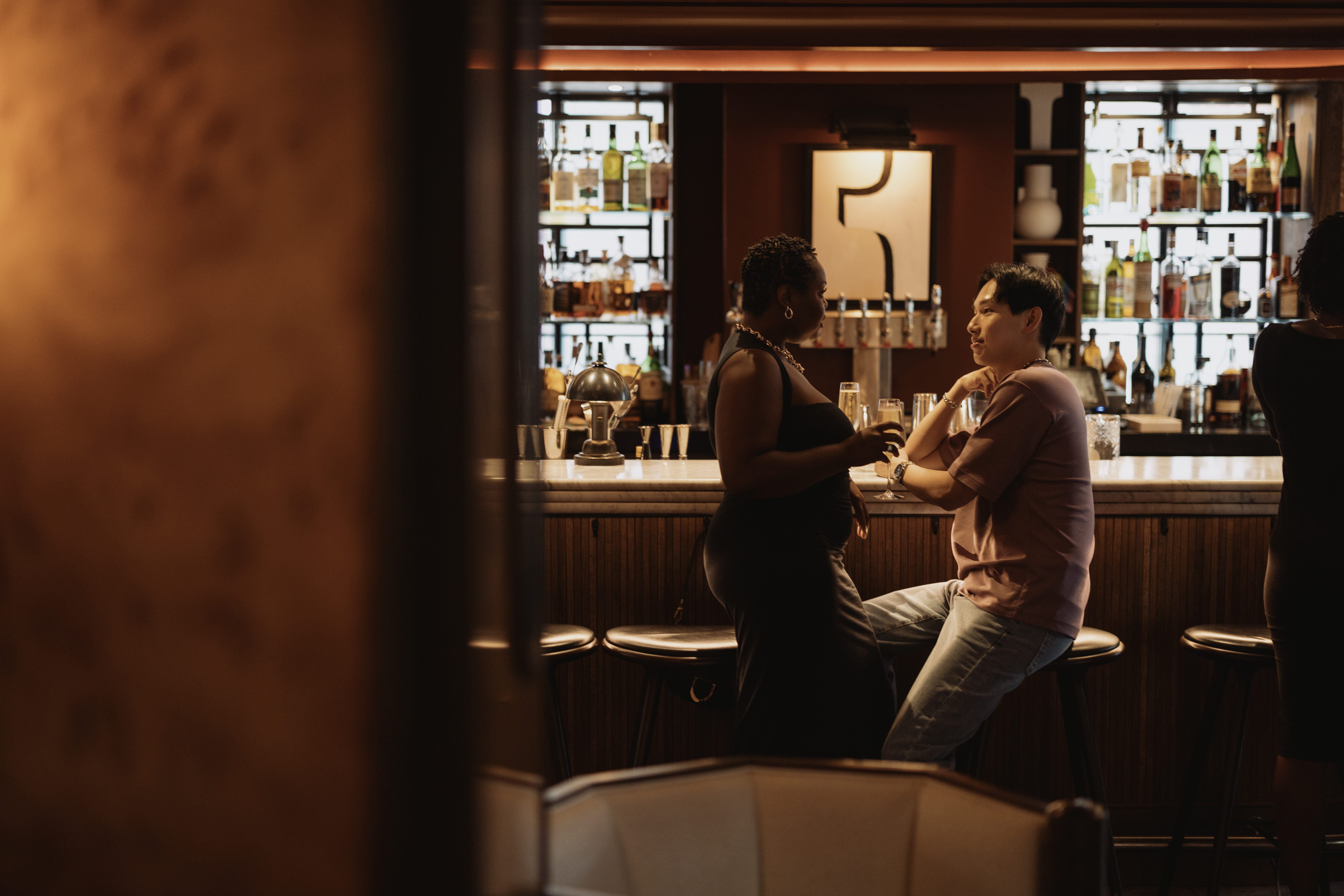 A couple enjoying drinks at a dimly lit bar in DC, engaged in conversation with shelves of liquor bottles in the background.